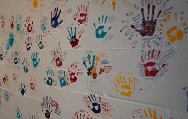Multicolor painted handprints on an otherwise blank concrete wall. Each handprint has a name and short message of a student next to it