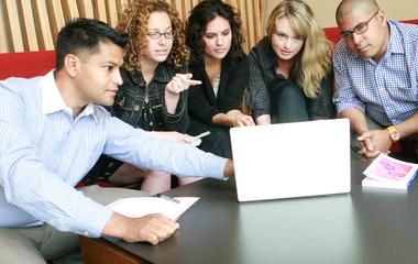 Five professionals engaged in a collaborative discussion around a laptop.