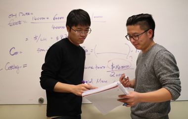 Two individuals discussing over documents with a whiteboard filled with notes in the background.