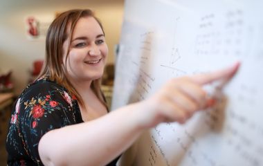 Woman pointing at a whiteboard with notes, smiling at the viewer.