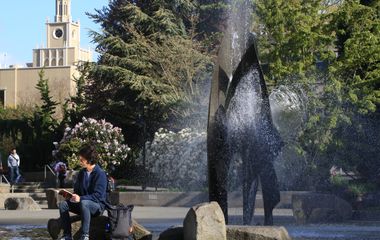 A person sitting near a water fountain on a sunny day with a building in the background.