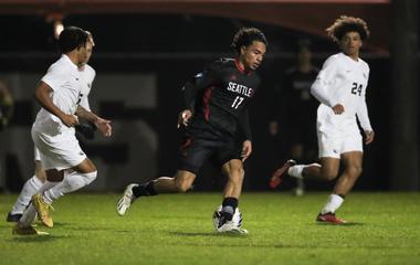 Three soccer players in a night match, with one controlling the ball and two others in pursuit.