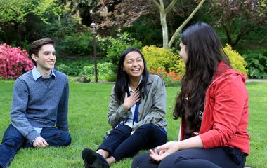 Three students laugh as they sit in a grassy field