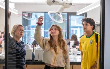 Photo of two students and professor writing formulas over a clear glass.