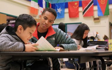 Teacher assisting a student with schoolwork in a classroom decorated with international flags.