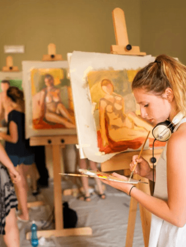 Woman looking down at paint palette while adding paint in front of a life painting