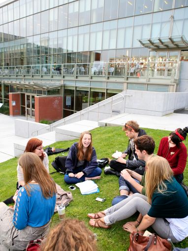 Students sitting outside the library on grass reading books and laughing in a circle.