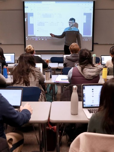 A classroom with a teacher in front of a projection screen and students in the foreground.