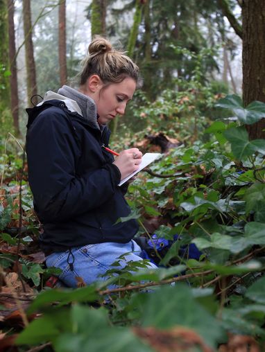 A student wearing fleece and a rainjacket kneeling in a forest writing in a notebook
