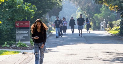 A woman walking towards the camera on a sunlit tree-lined path, checking her phone, with several people in the background walking away.