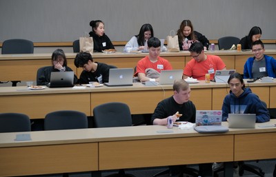 Students sit at desks with computers talking with one another