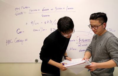 Two students look at a notebook as they stand in front of a whiteboard with writing on it