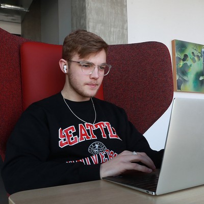 A computer science student in a big chair, wearing earbuds, and typing on a laptop computer.