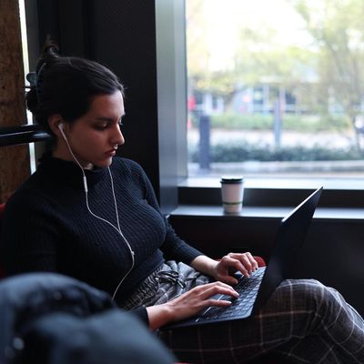 A student wearing earbuds and typing on a laptop computer with a coffee next to her