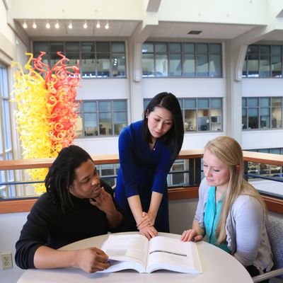 A professor and two students look at a page in a textbook at a round table with a glass Chihuly sculpture in the background