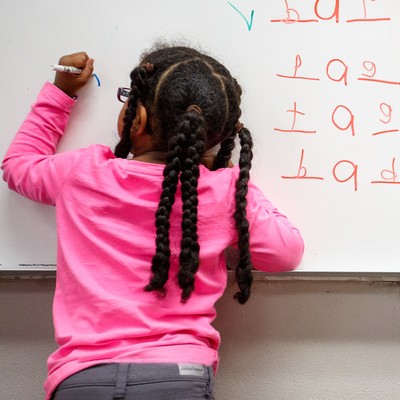 Student in pink shirt writing with left hand on whiteboard practicing spelling.