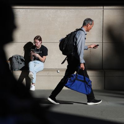Girl sitting outside on phone while man walks past her on his phone.