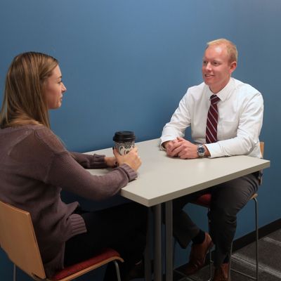 Two professionals engaged in a conversation at a small table in an office setting.