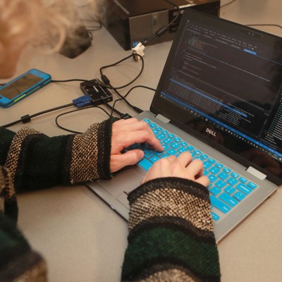A woman working on a laptop in a room with other people.