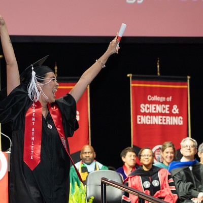 a student athlete in commencement regalia with arms raised in celebration