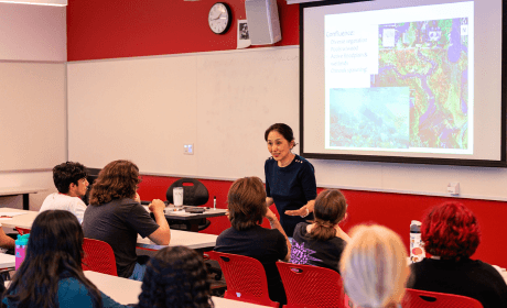 Professor stands at front of room and speaks to students sitting in chairs