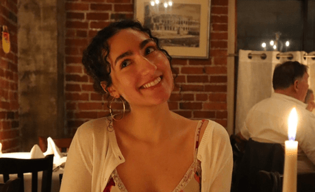 A woman sitting at a table with a drink smiling but looking away from the camera