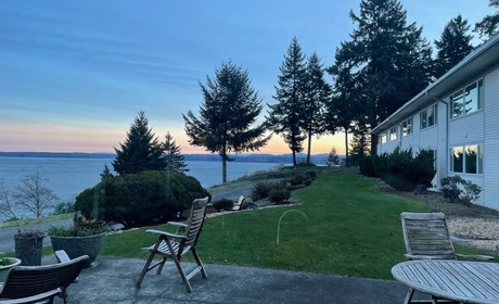 Photo of retreats center landscape. There is a lake on the left side, part of the building windows on the right side. A chair and grass are on the center. Green pines appear around. Above, a blue sky.