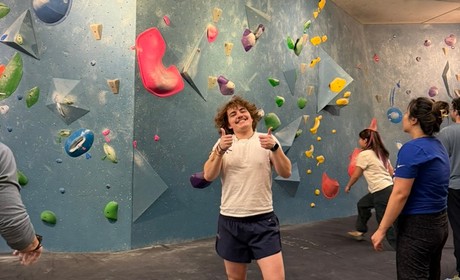 Photo of a student smiling. On the background, a blue climbing wall and some more students.