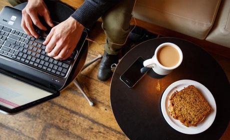 Hand typing on a laptop next to their coffee, pastry, and phone