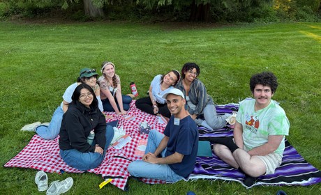 Photo of a group of seven students sitting on the grass over a picnic blanket. They are all smiling and some notebooks are over the blanket.