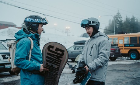 Two snowboarders standing in a snowy parking lot.