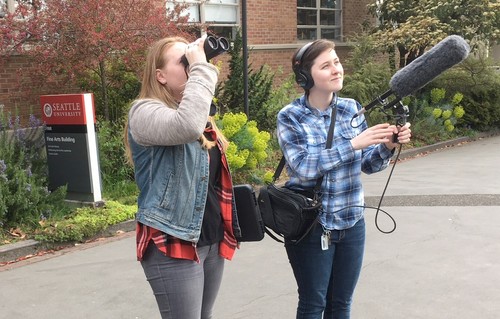 Two biology students, one with binoculars, the other with headphones, microphone, and an audio recorder, observing and audio recording birds.