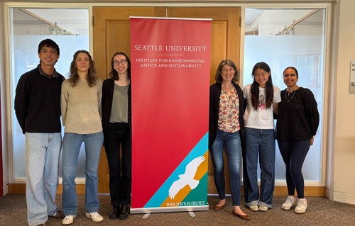 Six people standing and smiling next to a red banner reading 