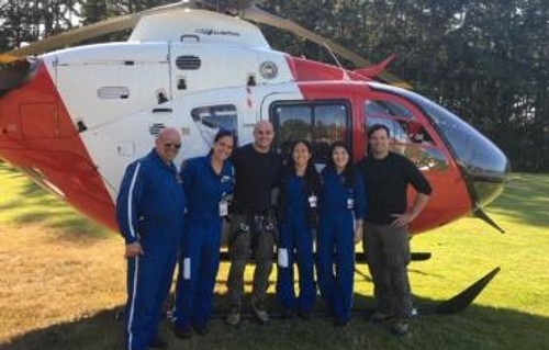 6 people standing in front of a red and white helicopter sitting on a field. 