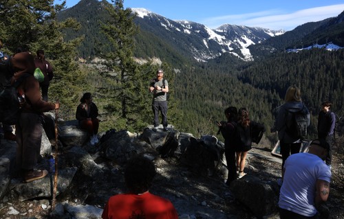 Dr. Eric Severson on the Iron Goat Trail, standing on a rock speaking with trees and mountains behind him