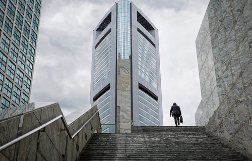 A small figures going up the steps toward a glass-enclosed building