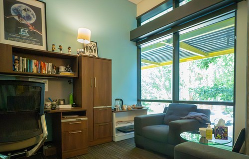 A picture of counselors office, with warm lighting, a movie poster, and decorations such as bobbleheads and coffee mugs, all in front of a large, bright floor to ceiling window.