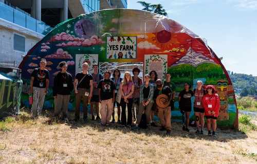 Group of students, Yes Farm staff, and faculty standing in front of colorful greenhouse with Yes Farm sign
