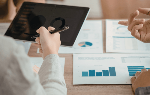 Two hands pointing at a tablet, in the background a table full of papers with multiple graphs and charts on them