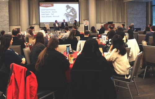A conference with the audience around tables listening to three people on stage.